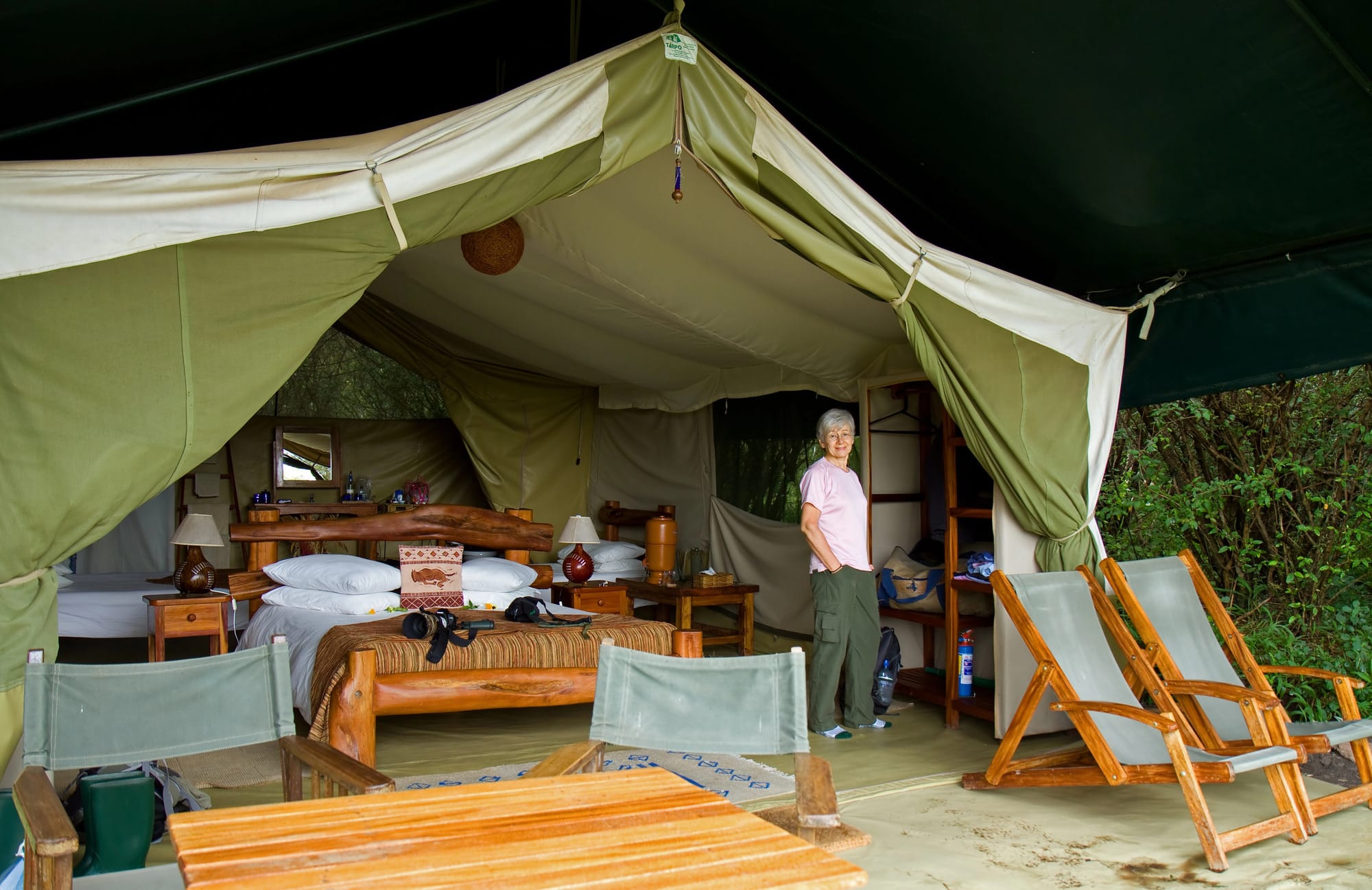 The inside of a safari tent, with a wooden bed, writing table and chairs on the veranda